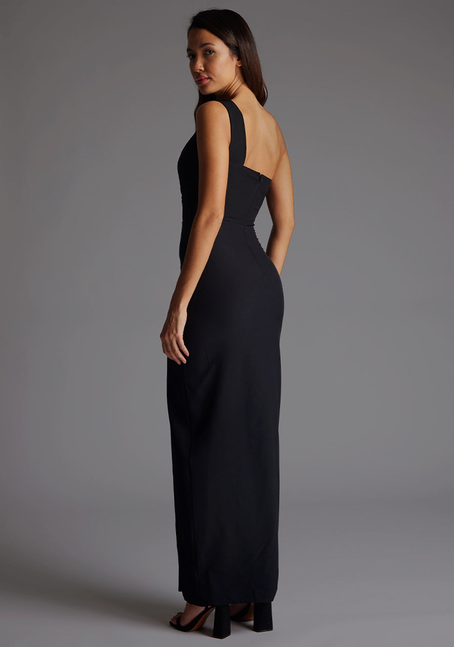 Side back image of the model wearing a black maxi dress with a sleek one-shoulder silhouette, contemporary front cut-out and front skirt split for effortless chic.  The design features Vesper Ellis Cut-Out One-Shoulder Maxi Dress in Black.