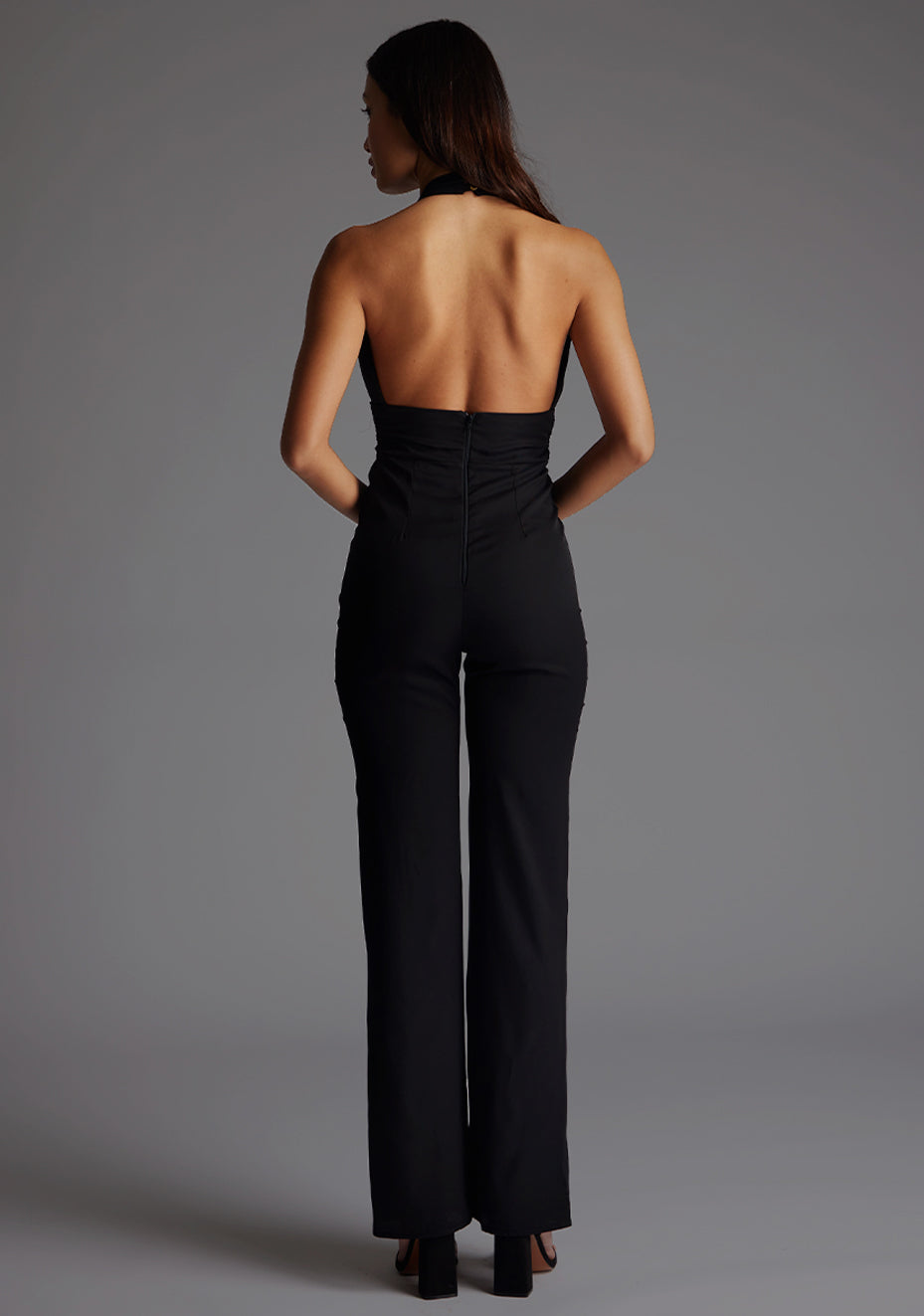 Back image of the model wearing a black jumpsuit with a chic plunge neckline and its open back adds a hint of allure. The design featured is the Vesper Flynn Plunge Neckline Wide Leg Jumpsuit in Black.
