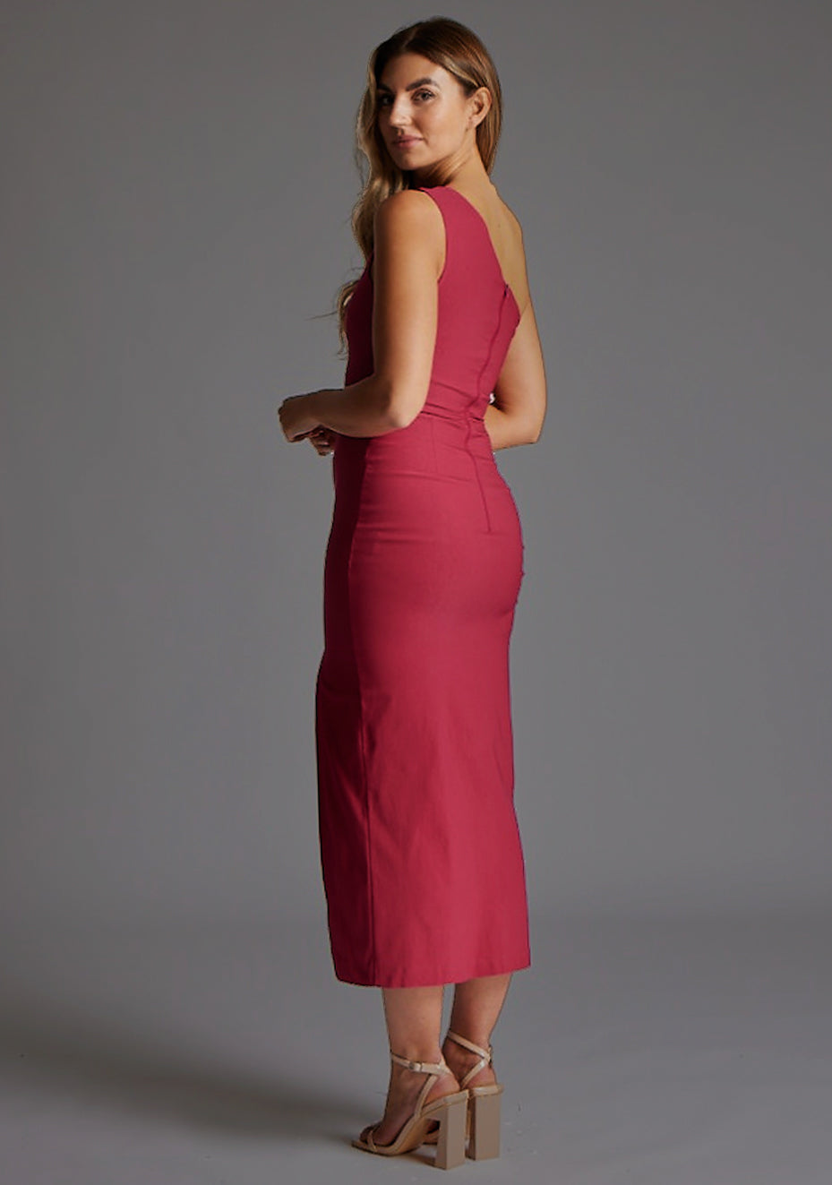 Side back image of blonde model wearing a raspberry one shoulder midaxi dress with a asymmetric neckline, one shoulder and front skirt split. The design featured is the Vesper Gianna One Shoulder Midaxi Dress in Raspberry