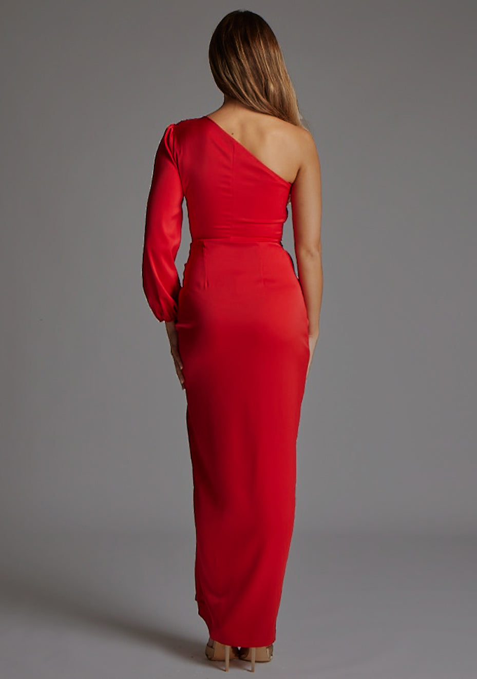 Back image of blonde model wearing a red one sleeve maxi dress with a asymmetric silhouette adds a touch of uniqueness, while the one long-sleeve design provides a sophisticated twist. The front cut-out detail adds a subtle allure. The design features Vesper Keaton One Sleeve Maxi Dress in Red.