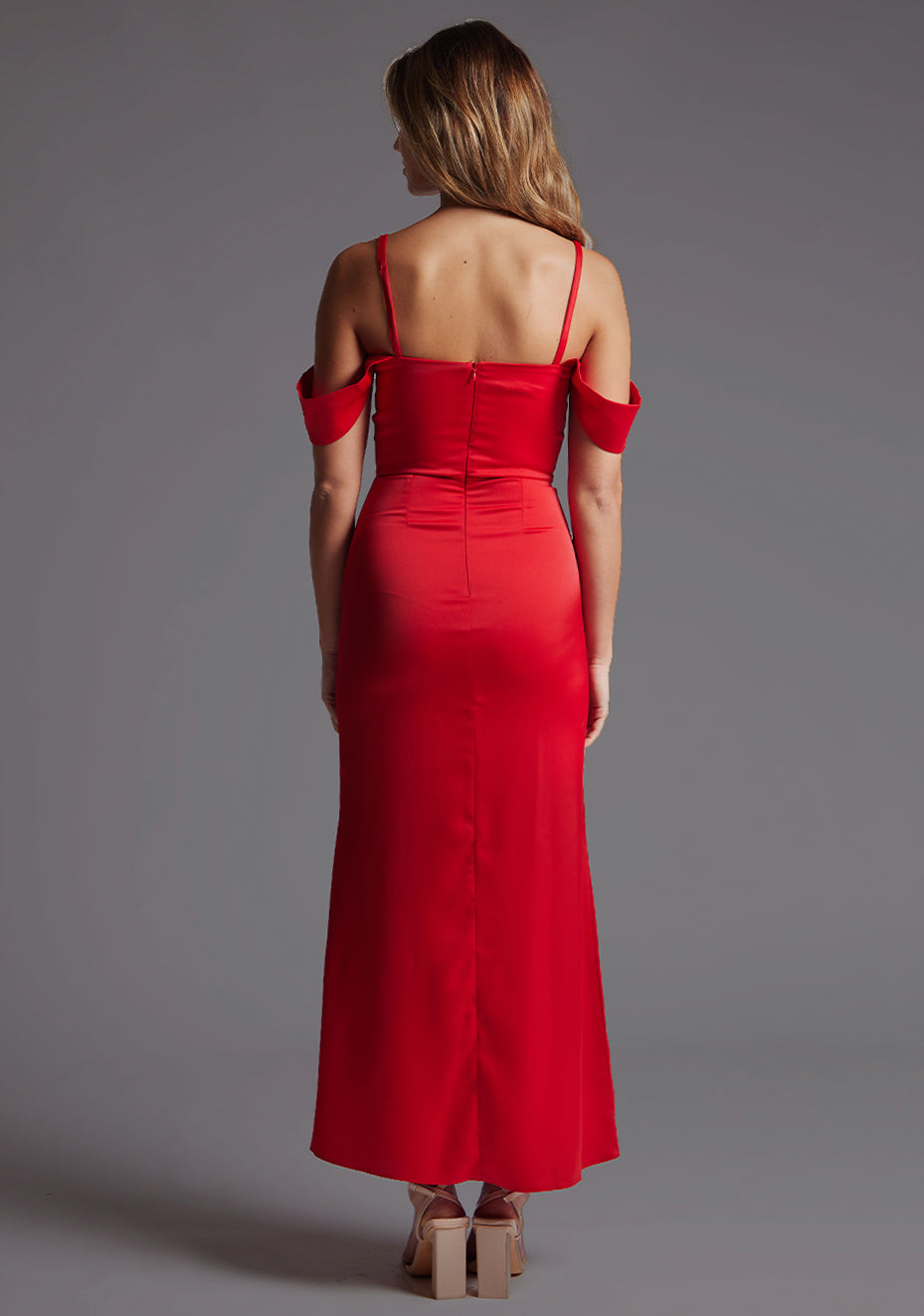 Back image of a blonde model wearing a red maxi dress with a striking black off-shoulder strap detail, beautifully complementing its slim-fit silhouette and zip closure at the back for ease and a seamless finish. The design featured Vesper Luisa Bardot Satin Maxi Dress in Red.
