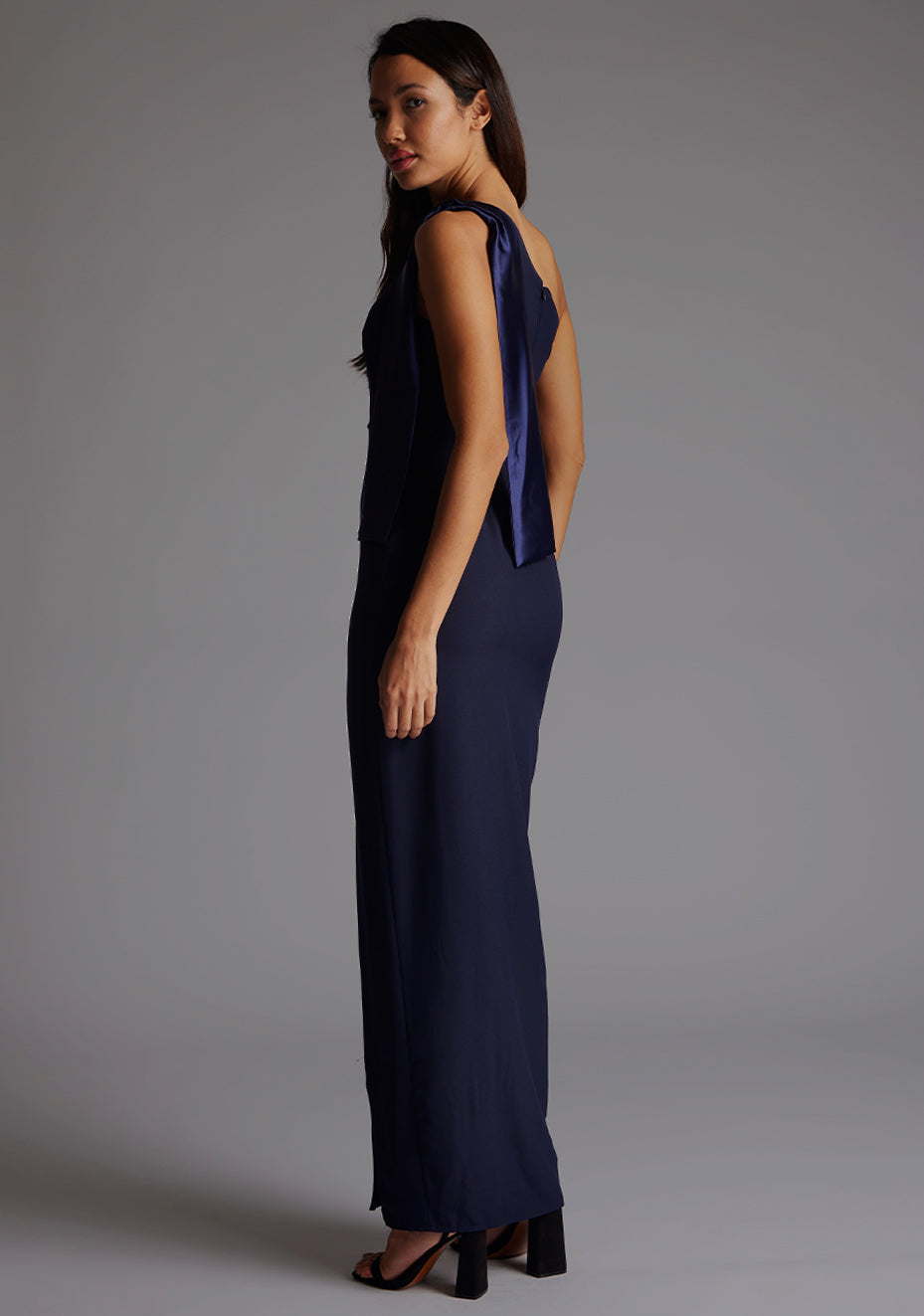 Side back image of a model wearing a  One Shoulder Navy Maxi Dress with a  one shoulder contrast satin band and bow detail with a front skirt split. The design features Vesper Olympia One Shoulder Maxi Dress in Navy.