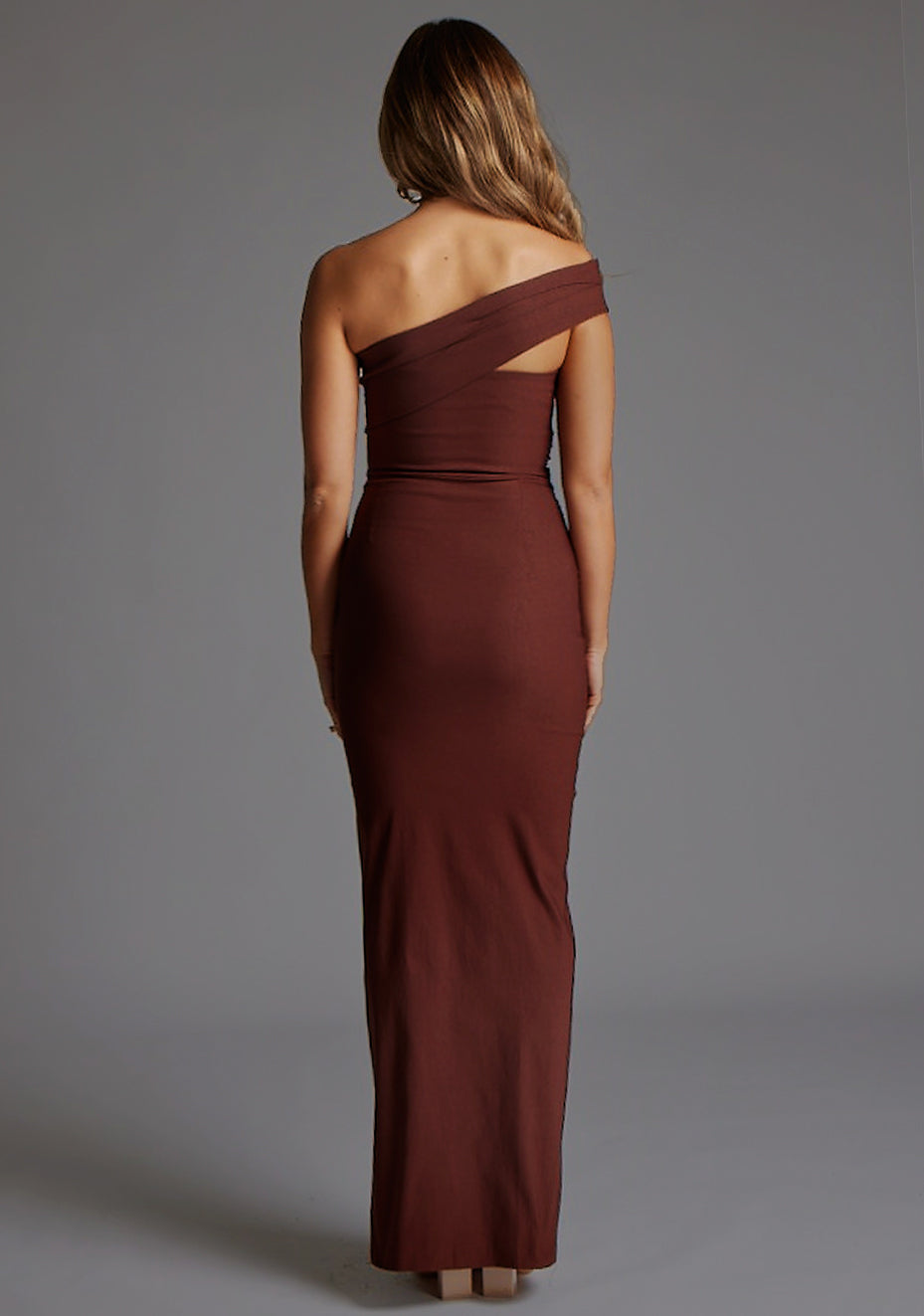 Back image of a blonde model wearing a  Chocolate Maxi Dress with an asymmetric neckline and one-sleeve design and front skirt split adds allure. The design features Vesper Pearson Bardot Ruched Maxi Dress in Chocolate.