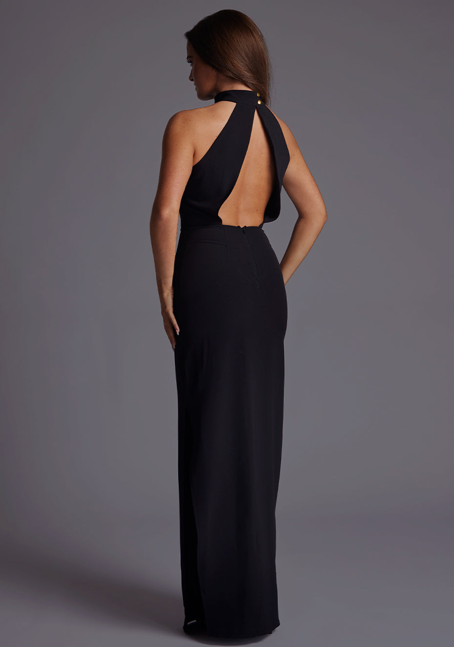Back quarter image of a model wearing a black cut out maxi dress with a elegant maxi dress features a split at the left side, open back and front bodice keyhole detail. This number fastens at the back of the neck with a buttoned collar and centre back zip detail. The design featured is the Vesper Kennedy Black Cut Out Maxi Dress
