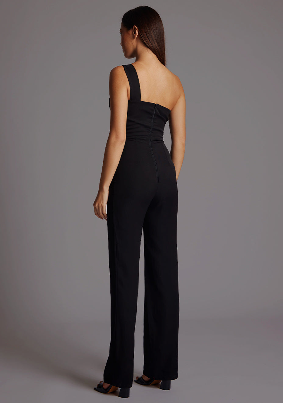 Back image of a  model wearing a black Jumpsuit with a stylish one-shoulder design, accentuated by a sophisticated front cut-out, slim fit and convenient back zip. The design featured is the Vesper Rosemary Black Jumpsuit.
