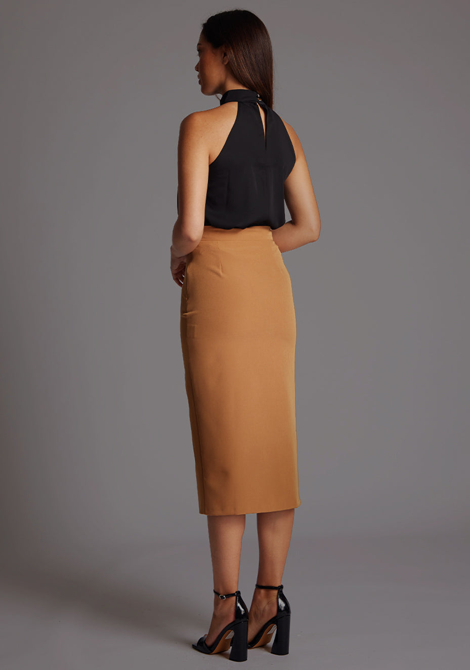 Side back image of a model wearing the Vesper Sherine High Waist Pencil Skirt in Camel, showing the smooth fit and clean lines from behind.