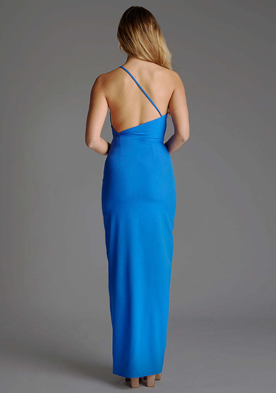 Back image of a model wearing a cobalt blue maxi dress with a asymmetric neckline, one shoulder strap, front skirt split and open back with asymmetric strap across back. The design featured is the Vesper Viva Cobalt One Shoulder Maxi Dress.
