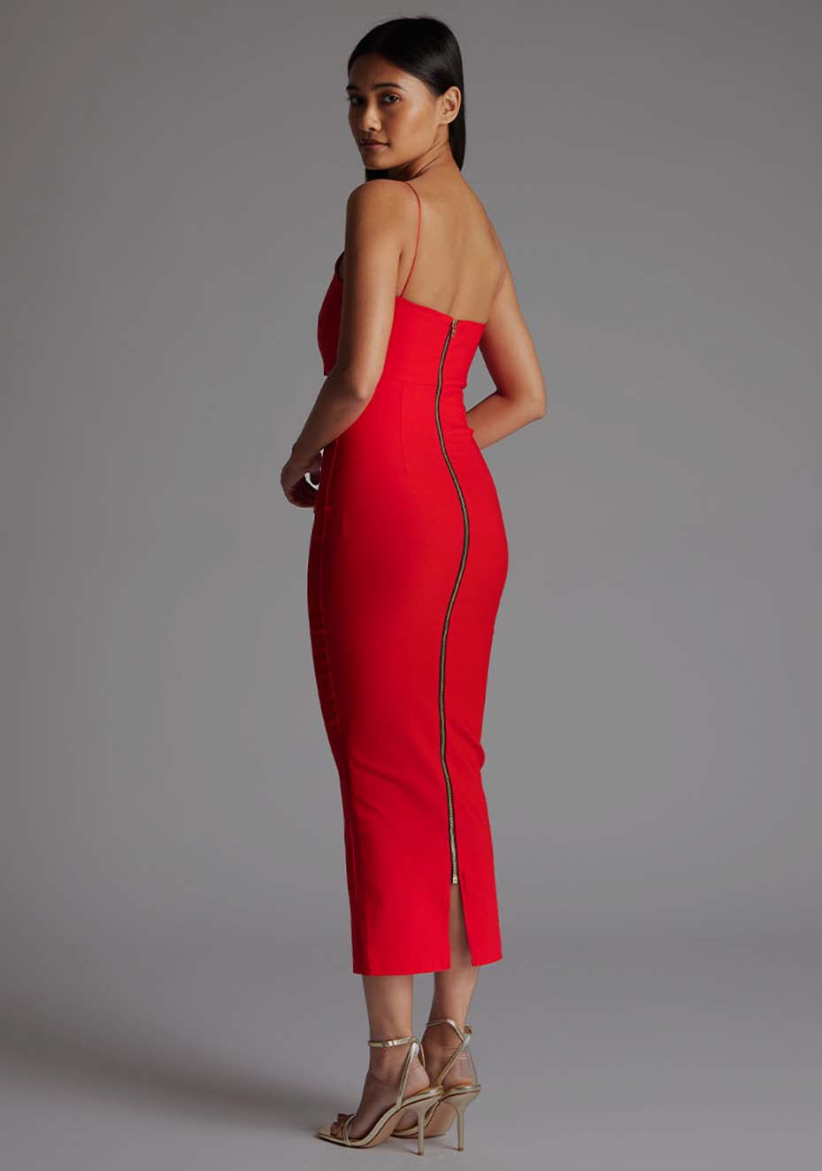 Back qaurter image of a model wearing a red midaxi dress with a asymmetric neckline and one shoulder with double thin strap detail on shoulder and across chest create a unique and elegant design, while the side waist cut-out and centre back gold zip add an extra touch of glamour. The design featured is Vesper Whitney Red Midaxi Dress
