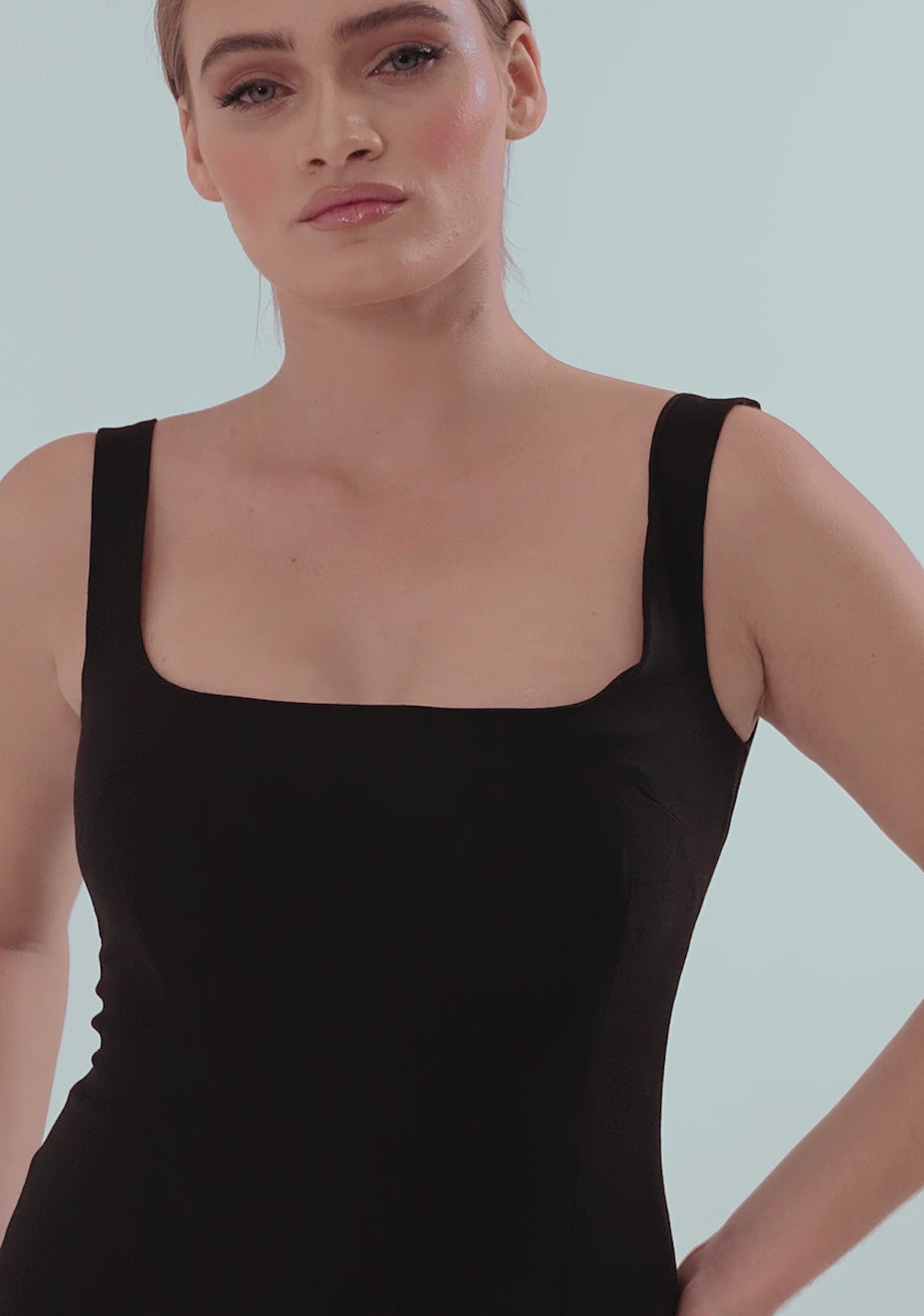 Model posing and turning in the VSPR Zada Black Midi Dress, showcasing the square neckline, body-hugging fit, and curve-enhancing silhouette in a sleek studio setting. The stretch luxe fabric subtly catches the light as she moves, emphasizing the elegant, minimal design of this classic little black dress.